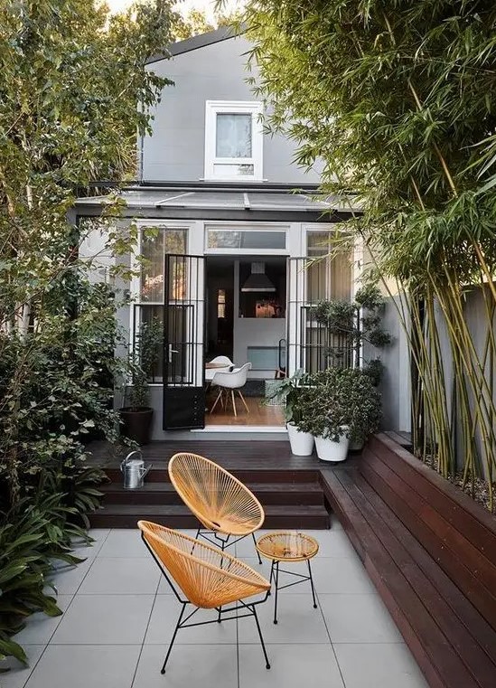 a small minimalist paved patio with a dark stained deck and a tiled floor, with growing bamboo and plants and chic woven chairs and a table