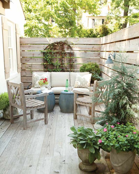 a small patio with a sofa, wooden chairs, mini side tables, potted greenery and blooms is a cozy rustic nook