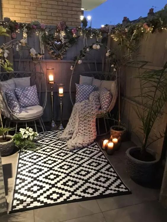 a small patio with egg-shaped chairs with pillows, printed pillows and a rug, potted plants and blooms, lights and candles