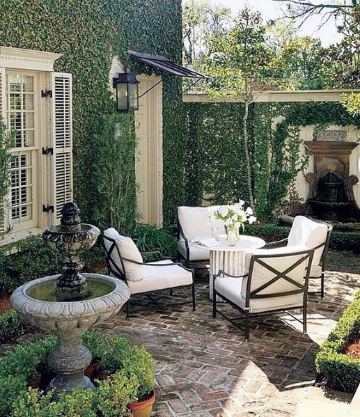 a small refined patio with a coffee table, white chairs, a fountain and lots of greenery, climbers on the walls of the house