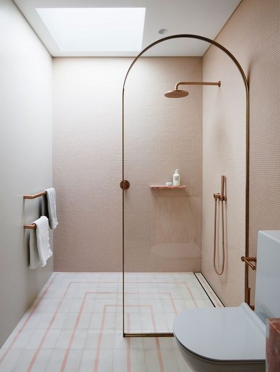 a small unique bathroom clad with blush mini tiles, with neutral and pink ones on the floor and a skylight over the shower plus brass touches