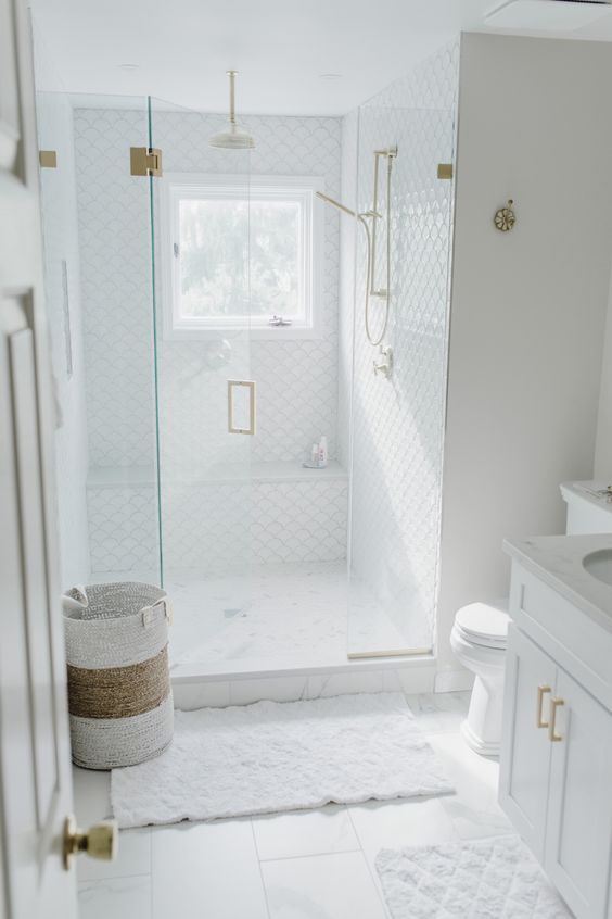 a small white bathroom with large scale and fish scale tiles, a shower space, a white vanity and a basket plus gold fixtures