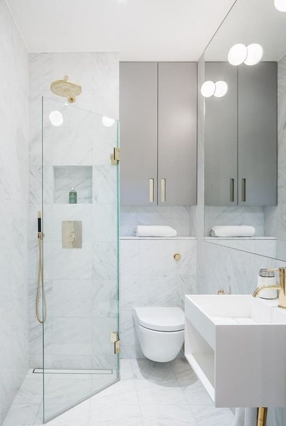 a tiny minimalist bathroom with white marble tiles, sleek grey cabinets, gilded touches and a triangle shower space