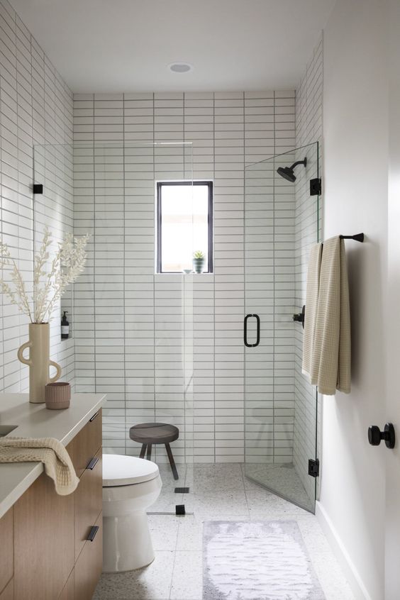 a tiny neutral bathroom with white skinny tiles and a shower space, a stained vanity and black fixtures plus a small window