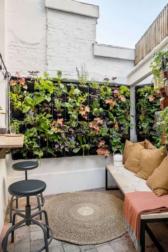 a tiny patio with a bench and pillows, a walk through window, black stools and gorgeous living walls