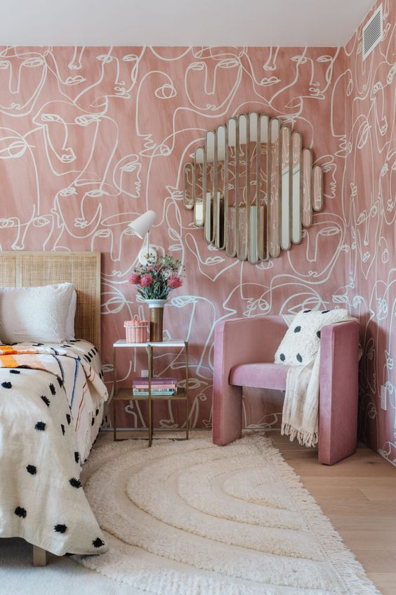 a whimsical pink bedroom with printed wallpaper, a bed with a cane headboard, a pink chair, a rug and a creative mirror