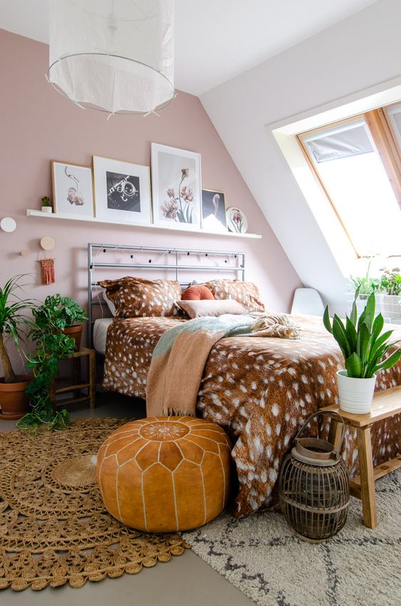 an attic pink boho bedroom with a ledge gallery wall, a metal bed with printed bedding, layered rugs, potted greenery and a lantern