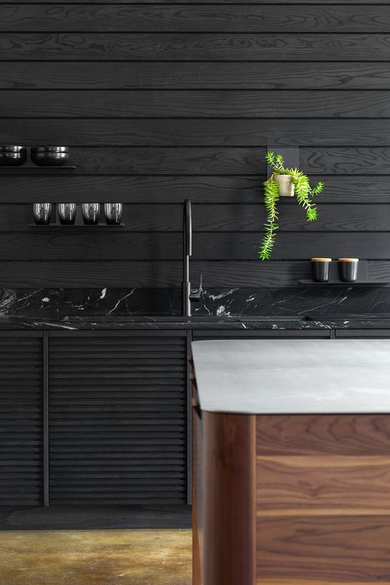 a moody kitchen with black plank walls, a black marble countertop and a wooden kitchen island with a white stone countertop