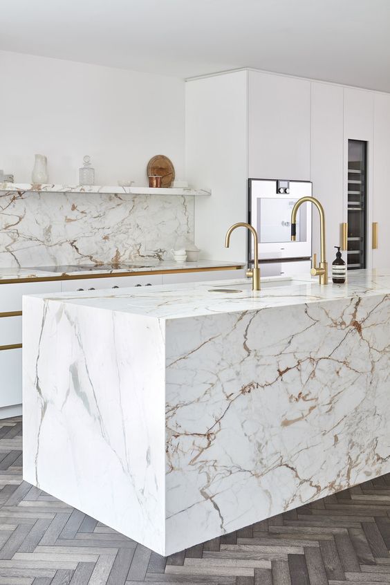 a modern luxurious kitchen with white cabinets, a white marble kitchen island and a backsplash plus gold fixtures