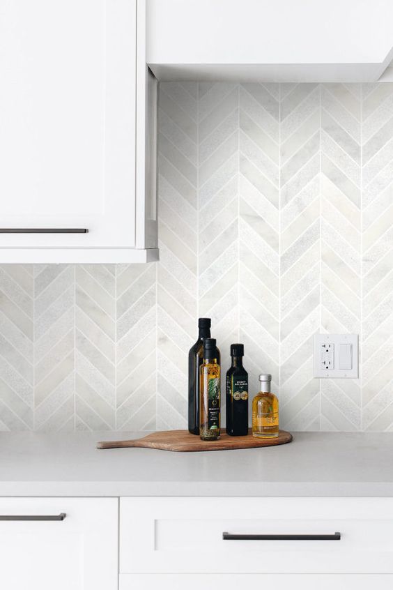 a white kitchen with grey stone countertops and a white marble tile herringbone backsplash for a pattern