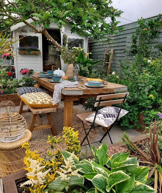 a bright and cool backyard with a lot of greenery, a wooden dining set and folding chairs, candle lanterns and blooms