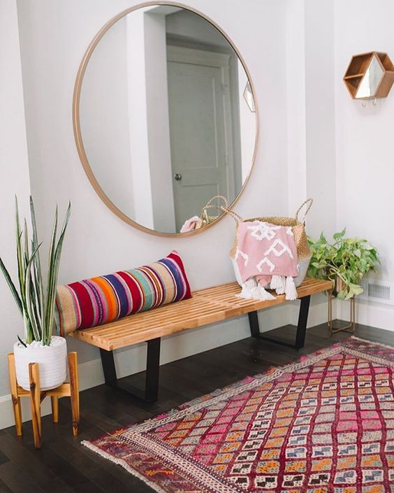 a colorful boho entryway with a wooden bench, a striped pillow, a round mirror, a bright rug and potted plants
