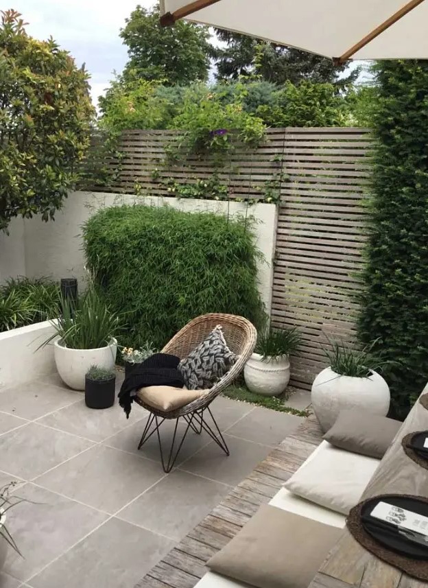 a contemporary walled backyard with a planked screen, with a tiled floor, potted grasses and greenery, a small dining set with pillows and a woven chair