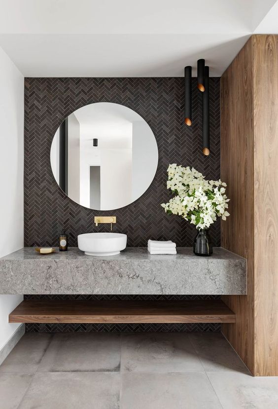 a gorgeous modern bathroom with a grey tile wall, black lamps, a grey stone vanity and a round mirror