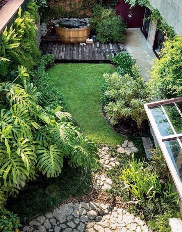a gorgeous small backyard with grass and lots of plants, with a desk with a wooden hot tub and a stone clad space
