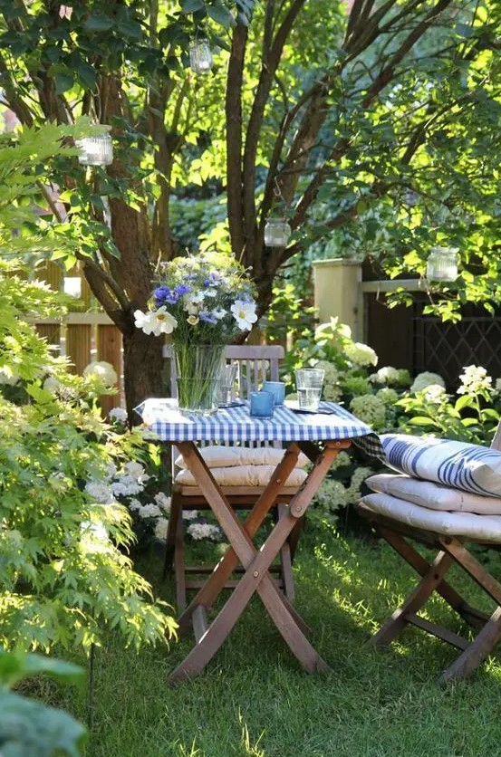 a little garden dining space on a green lawn, under the trees, with hanging candleholders and gold wooden chair plus cool textiles