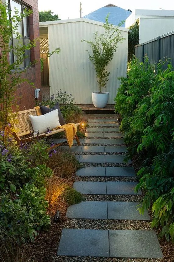 a long and narrow garden with a tile pathway, greenery, grasses and blooms, a rattan bench with pillows and some lights