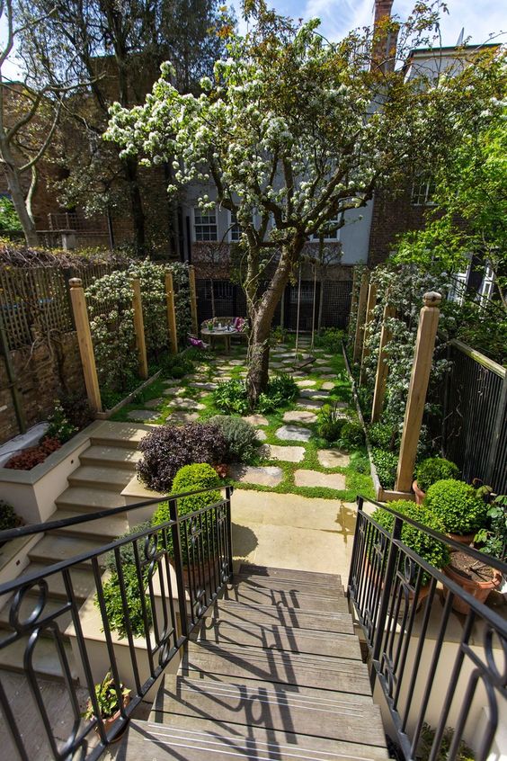 a lovely backyard garden with a blooming tree, greenery, a small table and a chair and potted greenery is amazing