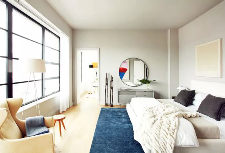 a lovely neutral modern bedroom with a comfy upholstered bed, a grey vnaity, comfy chairs and stools and a large round mirror