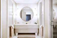 a neutral bathroom clad with white marble, with neutral walls and an oversized lit up round mirror to make the space even brighter