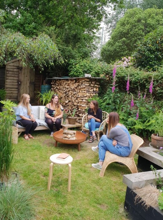 a rustic backyard with a green lawn with wooden and rattan furniture, with lots of greenery and bright blooms