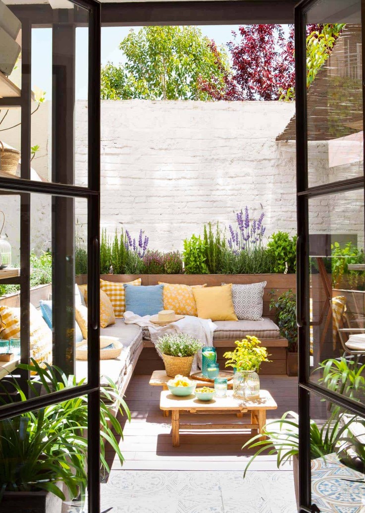 a small and bright patio with a corner sofa with lots of pillows, a couple of benches and blooms and greenery around