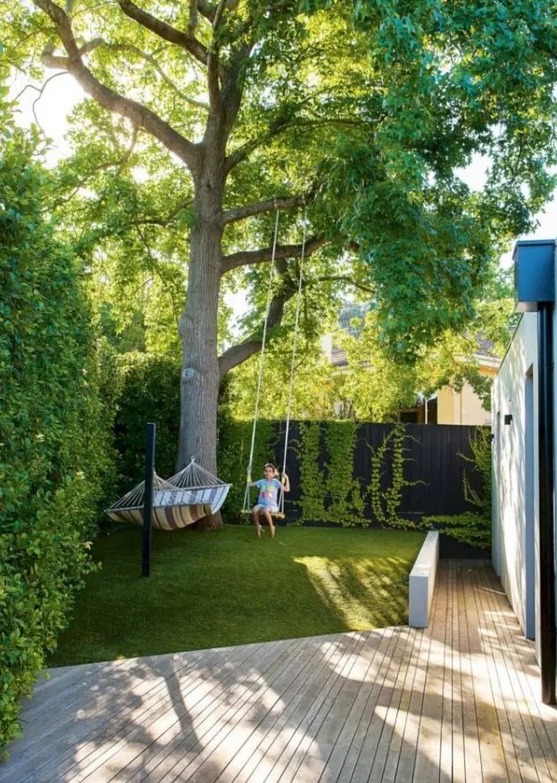 a small and dreamy backyard with living walls, a large tree, grass, a hammock and a swing, with a wooden deck