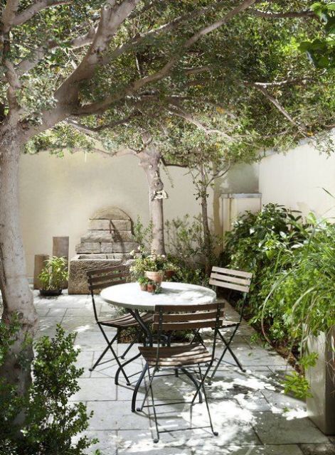 a small and elegant patio with lots of greenery, a stone deck, a small dining set, a tree and some wood decor