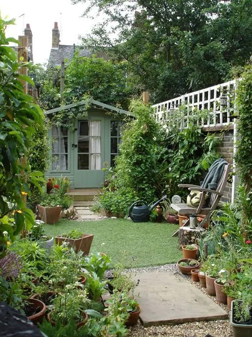 a small and narrow garden with a green lawn, greenery and shrubs, potted plants and a chair with a side table