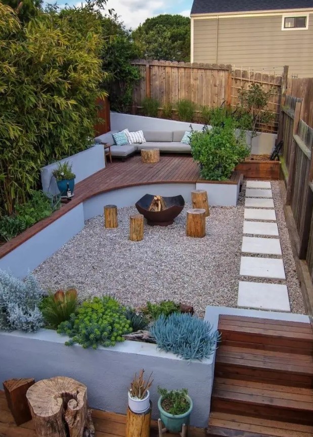a small and welcoming backyard with a fire pit zone and tree stumps with growing plants and trees, with a raised sitting zone with a sofa