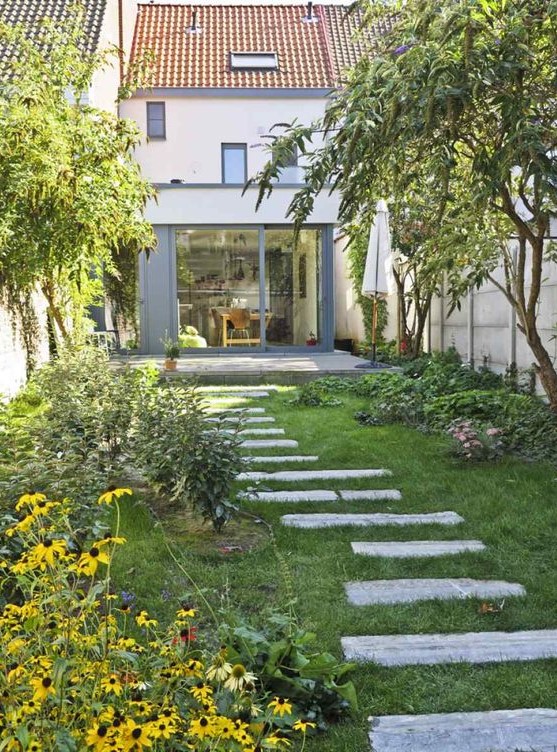 a small backyard garden with a green lawn, greenery and bright blooms and some trees plus a pavement