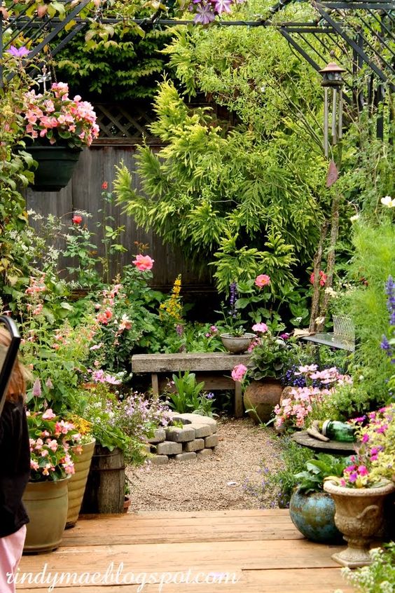 a small backyard turned into a garden with a stone bench, a fire pit and lots of greenery and blooms and trees around