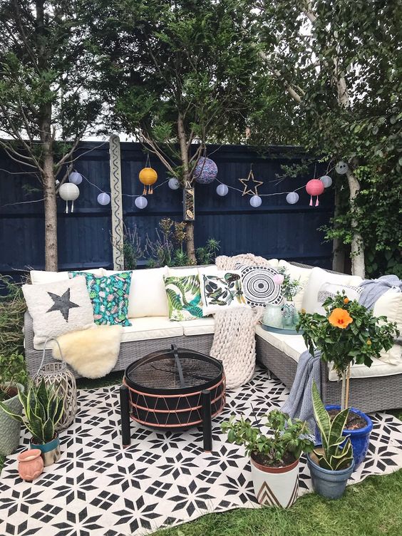 a small backyard with a green lawn, paper lamps, a wicker sofa with printed pillows and potted plants