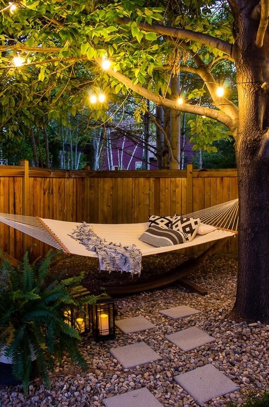 a small backyard with a hammock oasis, with some lanterns on the ground, ferns, string lights and printed pillows is a dreamy space
