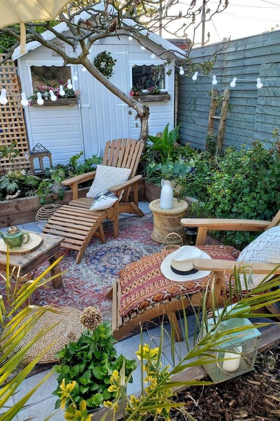 a small boho backyard with a boho rug, wooden loungers, some tables, greenery and blooms around, string lights and jute poufs