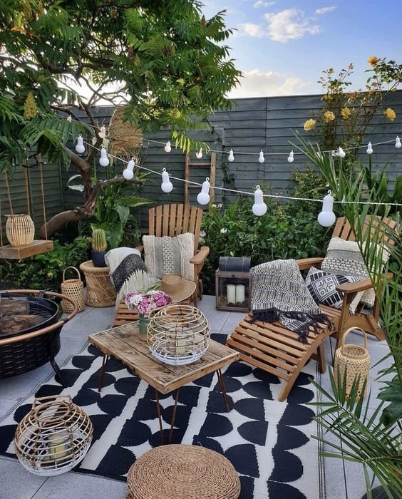 a small boho backyard with a wodoen deck, wooden loungers, a table, some jute poufs, candle lanterns, string lights and greenery