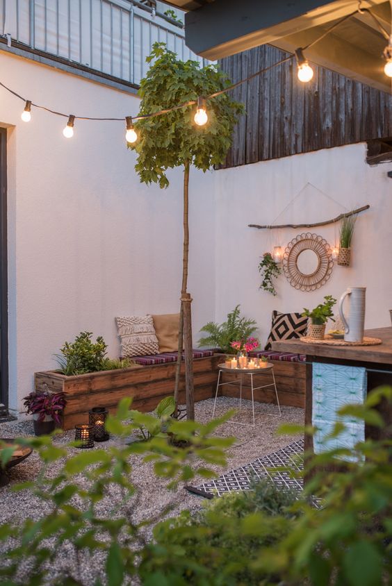 a small boho backyard with gravel on the ground, a wooden corner bench, some greenery, lights, boho pillows and lights