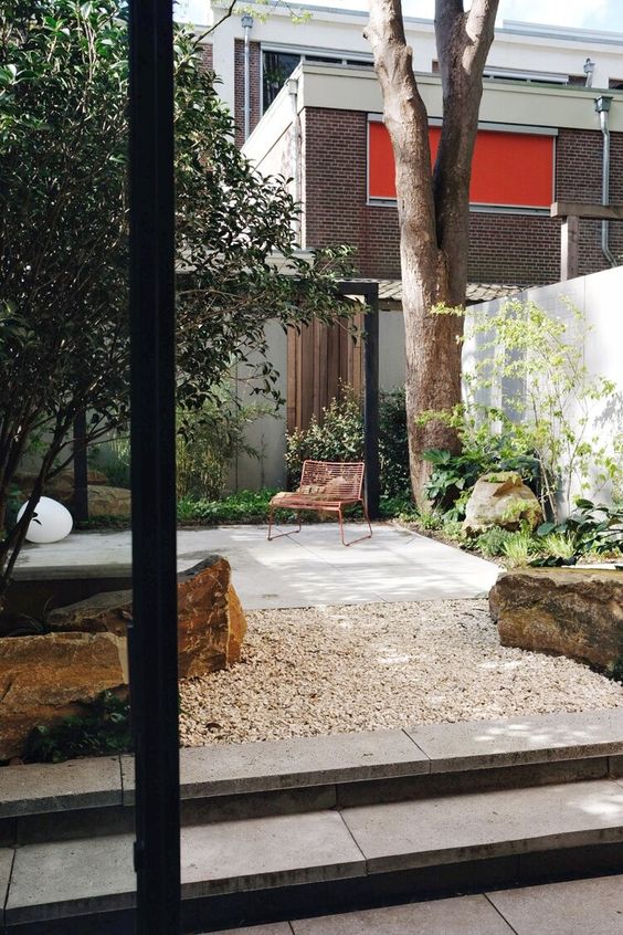 a small contemporary patio with a stone deck, some trees, greenery and plants, large rocks and pebble lamps on the ground