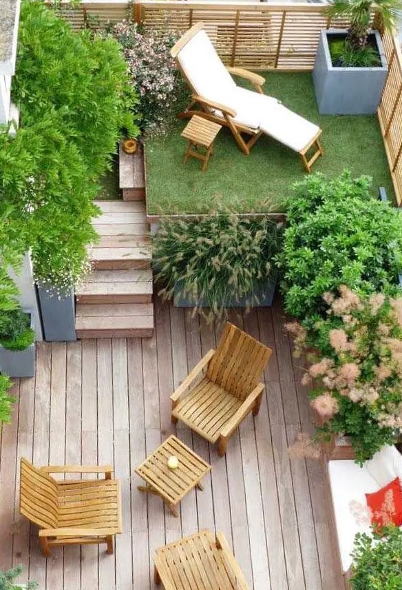 a small modern backyard with a raised platform with grass and a lounger, with potted trees, a ladder and a wooden deck with some wooden furniture