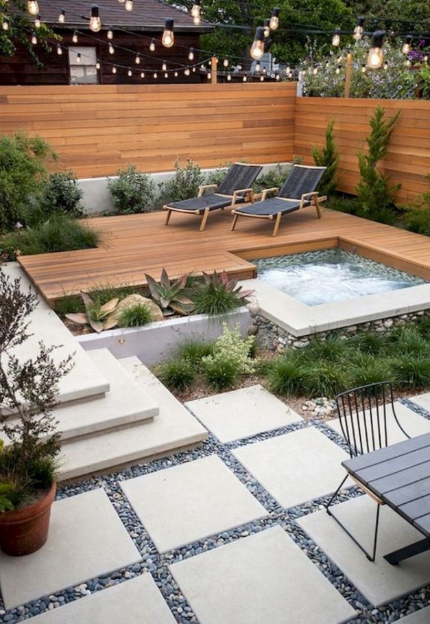a small modern backyard with a stained fence and deck, with a hot tub, growing plants and loungers, with a dinign table and chairs