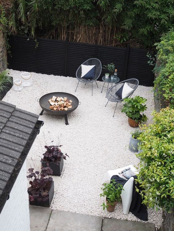a small modern backyard with white gravel, rattan chairs, a fire pit and some potted plants is super stylish