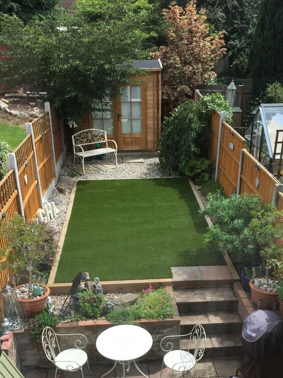 a small raised outdoor space with a green lawn, gravel around, some greenery and blooms and a bench plus a dining set
