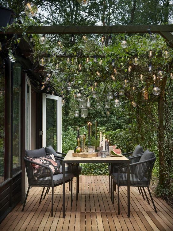 a small terrace with a wooden deci, a table and black chairs, beams with greenery and lights over the space