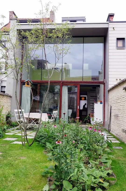 a small walled modern garden with a lawn, a tree, some blooms and greenery and a white dining set is very simple and cute