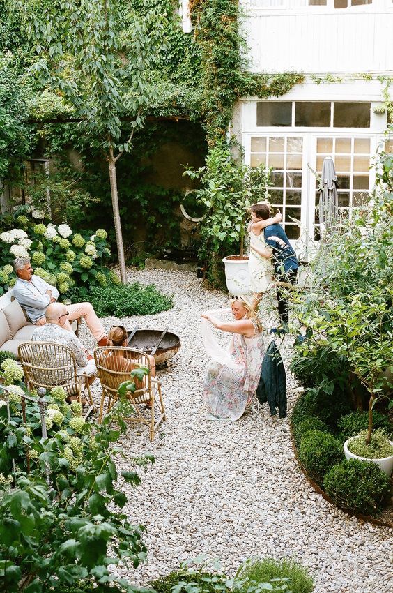 a small yet cozy backyard with gravel on the ground, some rattan furniture and lots of greenery and white blooms around