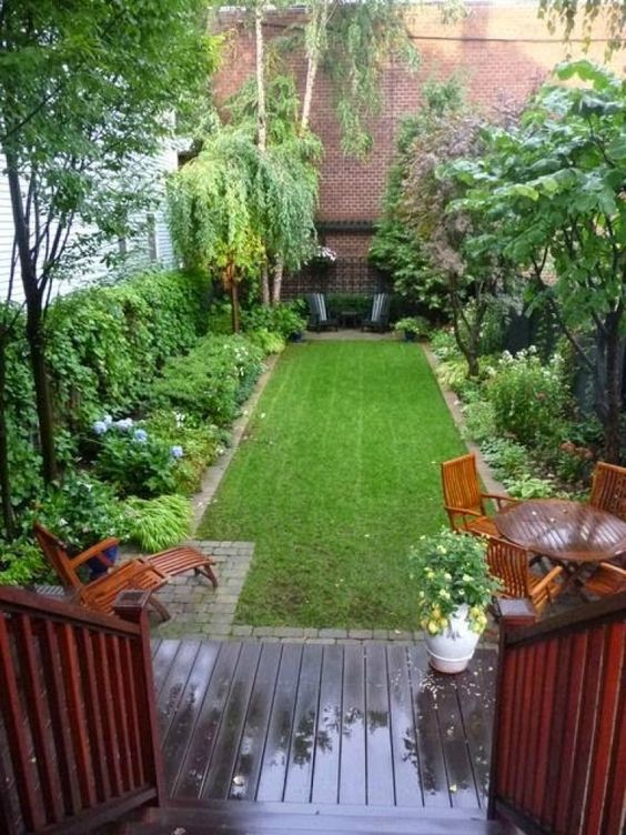 a small yet lovely backyard with a green lawn, some greenery and blooms around, a wooden dining set and a lounger and a wooden deck