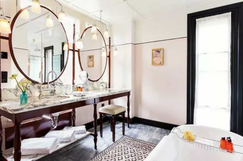 an eclectic bathroom with two oversized round mirrors, a long vintage vanity with a marble tabletop and a printed rug