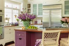 a neutral vintage farmhouse kitchen with a purple kitchen island, skylights and pendant lamps and some purple blooms