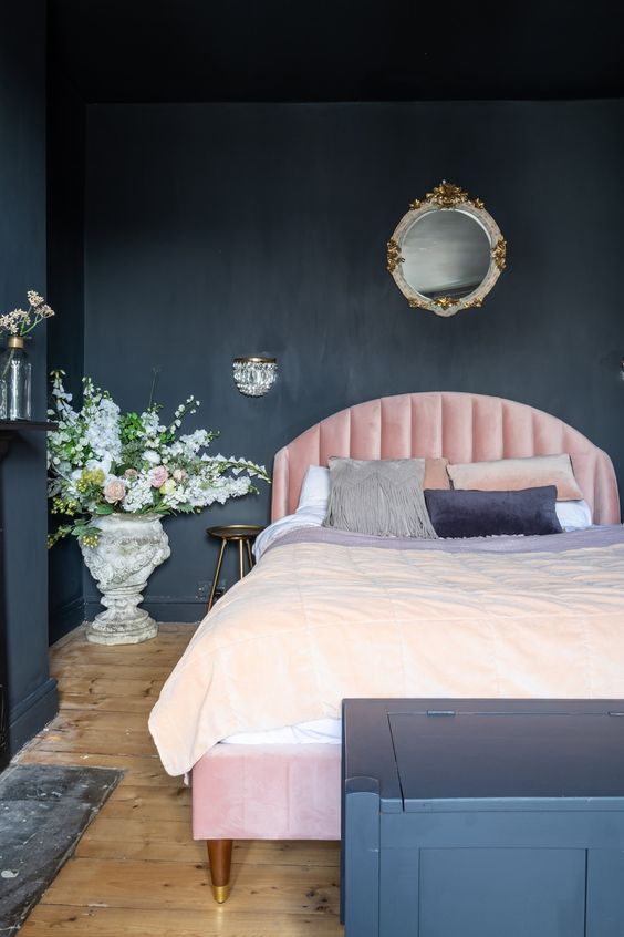 a chic moody bedroom with black walls and a chest for storage, a light pink bed, refined lighting and blooms