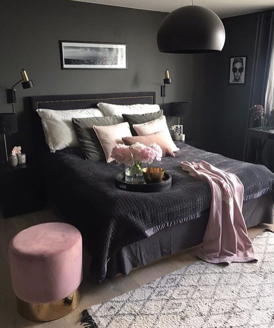 a moody bedroom with grey walls, a black bed, nightstand and a pendant lamp, pink and black bedding and a pink ottoman